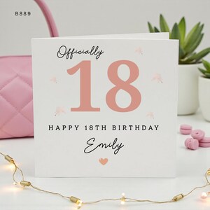 May include: A square white birthday card featuring the text Officially 18 in a large rose gold font. Below this, the text reads HAPPY 18TH BIRTHDAY followed by the name Emily in a black script font and a small rose gold heart icon.