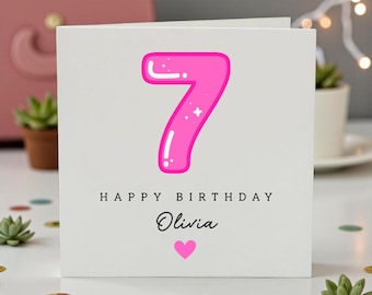 Op de afbeelding: Een vierkante witte wenskaart met een groot, glanzend roze cijfer 7 met witte accenten. Onder het cijfer staat de tekst HAPPY BIRTHDAY in zwarte hoofdletters, gevolgd door de naam Olivia in zwarte schrijfletters en een klein roze hartje.