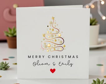 May include: A square white greeting card featuring a stylised gold Christmas tree graphic made of swirling lines and ornaments. The text reads MERRY CHRISTMAS followed by the names Oliver & Emily and a small red heart symbol. Code RSD 3094.