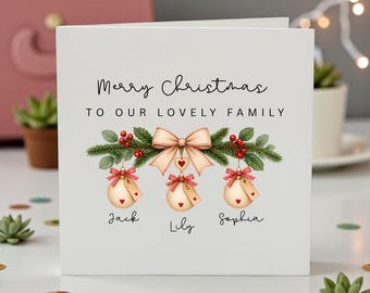 Pode incluir: Um cartão de felicitações de Natal quadrado branco com o texto Merry Christmas TO OUR LOVELY FAMILY. O design inclui um ramo de pinheiro verde com bagas vermelhas, um laço bege e três ornamentos pendurados com os nomes Jack, Lily e Sophia.