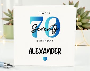 May include: A white birthday card with the words "Happy 70th Birthday Alexander" in black and blue. The number 70 is in blue, with the word "Seventy" in cursive. A blue heart is at the bottom of the card.
