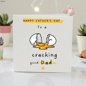 Puede incluir: Una tarjeta blanca del Día del Padre con el texto "Happy Father's Day" y "To a cracking good Dad". La tarjeta presenta una ilustración de un huevo roto con una yema amarilla.