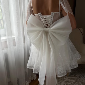 May include: White bridal dress with a large bow detail on the back. The dress features a corset-style bodice with lace-up closure and a short, flared skirt. Delicate tulle accents the shoulders and skirt, creating a romantic look.