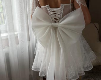 May include: White bridal dress with a large bow detail on the back. The dress features a corset-style bodice with lace-up closure and a short, flared skirt. Delicate tulle accents the shoulders and skirt, creating a romantic look.
