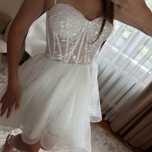 May include: A white, short dress with a sequined bodice and a layered tulle skirt. The dress features thin spaghetti straps and a corset-style top. The skirt is short and flared, creating a voluminous silhouette. The dress is designed for a formal occasion.