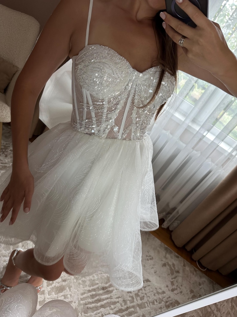 May include: A white, short dress with a sequined bodice and a layered tulle skirt. The dress features thin spaghetti straps and a corset-style top. The skirt is short and flared, creating a voluminous silhouette. The dress is designed for a formal occasion.