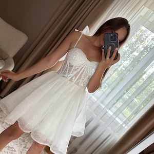 May include: A white, short, sparkly dress with a sweetheart neckline and spaghetti straps. The bodice is embellished with sequins, and the skirt is layered with tulle. The dress is designed for a formal occasion.