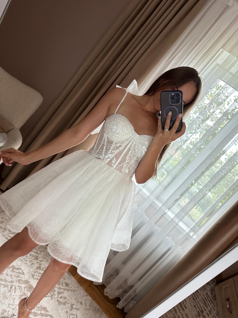 May include: A white, short, sparkly dress with a sweetheart neckline and spaghetti straps. The bodice is embellished with sequins, and the skirt is layered with tulle. The dress is designed for a formal occasion.