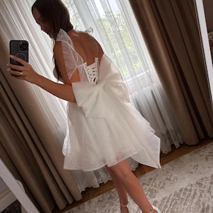 May include: A white, short dress with a layered skirt and a large bow on the back. The dress has a corset-style bodice and sheer, ruffled sleeves. The person is wearing white high-heeled sandals. The background includes curtains and a mirror.