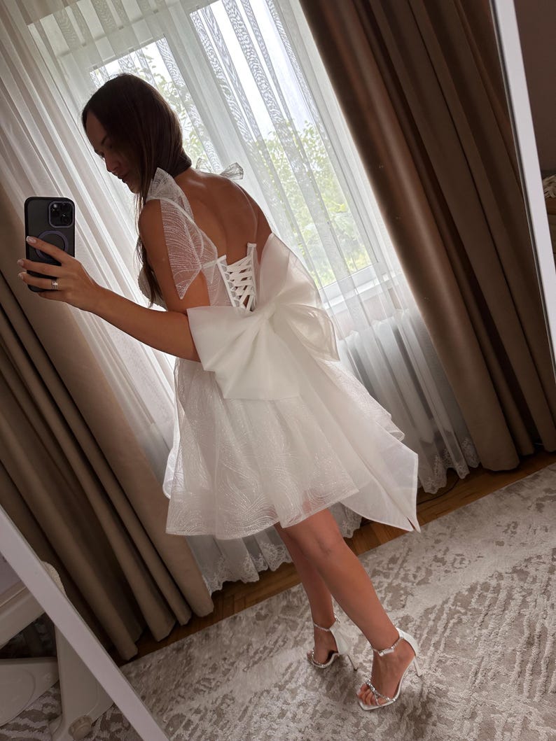 May include: A white, short dress with a layered skirt and a large bow on the back. The dress has a corset-style bodice and sheer, ruffled sleeves. The person is wearing white high-heeled sandals. The background includes curtains and a mirror.