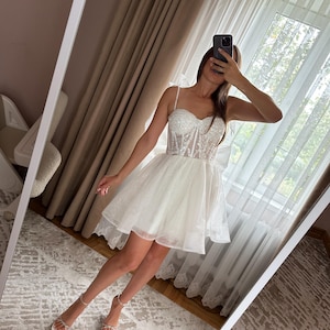 May include: White mini dress with a fitted, embellished bodice and a layered, flared skirt. The dress features thin straps and is paired with white sandals. The image is taken in front of a mirror.