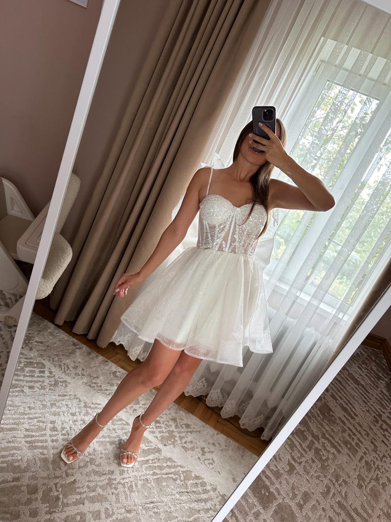 May include: White mini dress with a fitted, embellished bodice and a layered, flared skirt. The dress features thin straps and is paired with white sandals. The image is taken in front of a mirror.