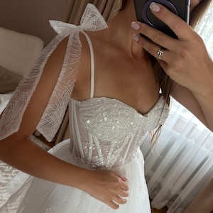 May include: A white wedding dress with a sweetheart neckline and a sparkling bodice. The dress features delicate spaghetti straps and sheer, bow-tied sleeves. The skirt is layered and flowing, creating a romantic and elegant look.