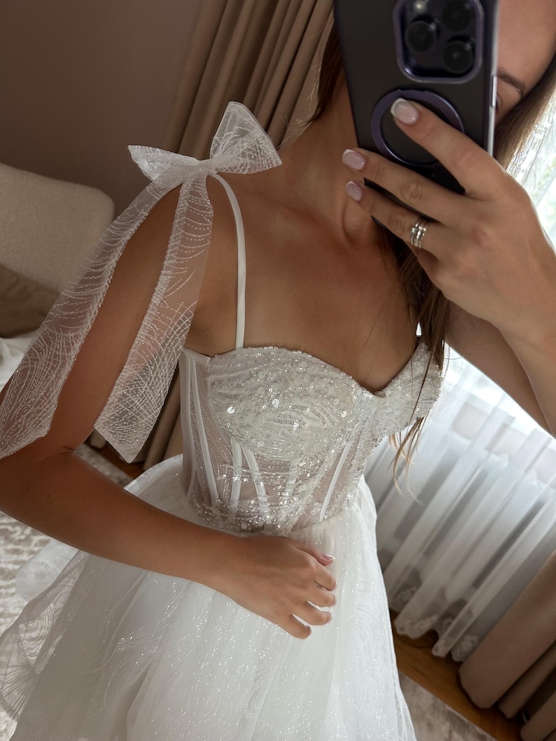 May include: A white wedding dress with a sweetheart neckline and a sparkling bodice. The dress features delicate spaghetti straps and sheer, bow-tied sleeves. The skirt is layered and flowing, creating a romantic and elegant look.
