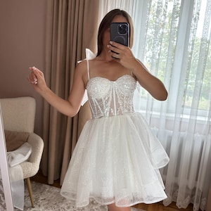 May include: A white, short, sparkly dress with a sweetheart neckline and spaghetti straps. The bodice is embellished with sequins, and the skirt is layered and flared. The dress is designed for a special occasion.