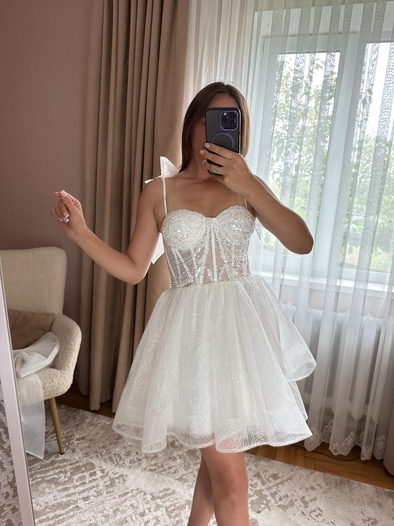 May include: A white, short, sparkly dress with a sweetheart neckline and spaghetti straps. The bodice is embellished with sequins, and the skirt is layered and flared. The dress is designed for a special occasion.