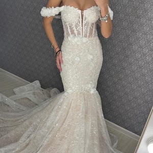 May include: Off-the-shoulder ivory wedding gown with floral embellishments and a mermaid silhouette. The dress features a sheer bodice and a long, flowing train. The gown is adorned with sequins, adding a touch of sparkle.