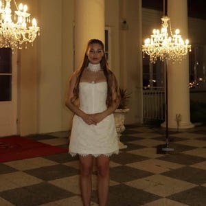 K&ouml;nnte beinhalten: Eine Frau in einem wei&szlig;en Kleid steht vor einem Geb&auml;ude mit gro&szlig;en S&auml;ulen und Kronleuchtern. Das Kleid ist kurz, mit Spitzenbesatz und passendem Choker. Sie tr&auml;gt wei&szlig;e Abs&auml;tze.