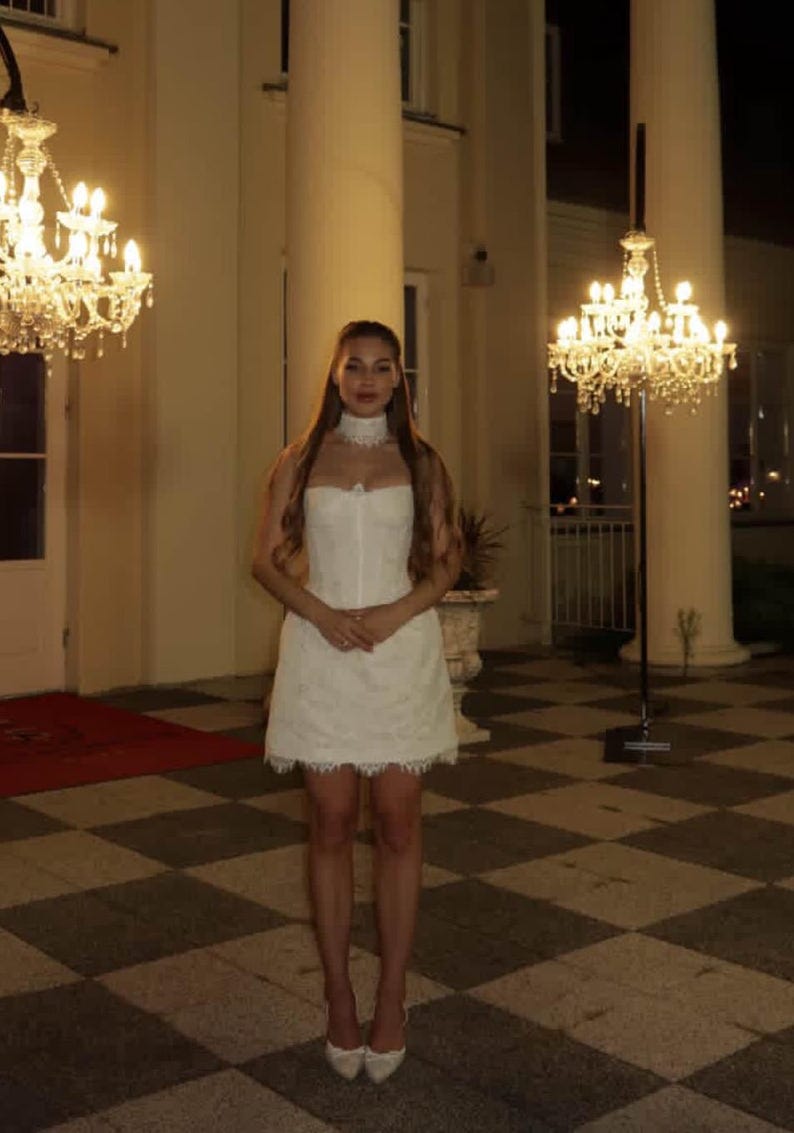 K&ouml;nnte beinhalten: Eine Frau in einem wei&szlig;en Kleid steht vor einem Geb&auml;ude mit gro&szlig;en S&auml;ulen und Kronleuchtern. Das Kleid ist kurz, mit Spitzenbesatz und passendem Choker. Sie tr&auml;gt wei&szlig;e Abs&auml;tze.