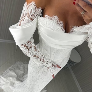 May include: A white floor-length gown featuring an off-the-shoulder neckline with a smooth satin band and long, fitted lace sleeves. The bodice and skirt are constructed from intricate floral lace with scalloped edges along the neckline and sleeves.