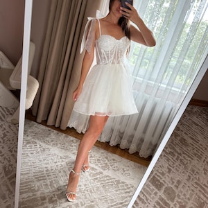 May include: A white, short wedding dress with a sweetheart neckline and a tulle skirt. The bodice is embellished with sequins and beads. The dress has delicate bow straps and is paired with strappy heels.