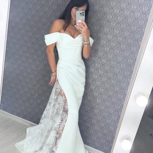 Könnte beinhalten: Ein bodenlanges weißes Kleid mit schulterfreiem Herz-Ausschnitt und figurbetontem Oberteil. Der Rock hat einen hohen Seitenschlitz, der einen transparenten Einsatz mit aufwendiger weißer Blumen-Spitzenstickerei bis zum Boden freigibt.