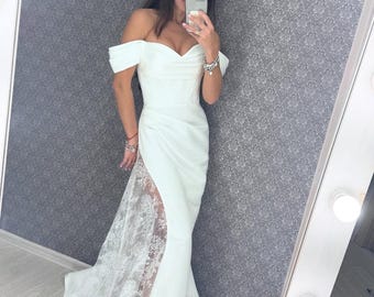May include: A floor-length white gown featuring an off-the-shoulder sweetheart neckline and a fitted bodice. The skirt includes a high side slit revealing a sheer panel with intricate white floral lace embroidery that extends to the floor.