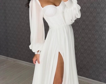 May include: A floor-length white dress featuring a structured corset-style bodice with a sweetheart neckline and long, sheer, voluminous bishop sleeves. The skirt is made of a flowing fabric and includes a high side slit that extends to the thigh.
