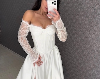 May include: A white wedding dress with an off-the-shoulder design. The bodice is fitted, and the sleeves are made of lace. The skirt has a high slit. The dress is made of a satin-like material.
