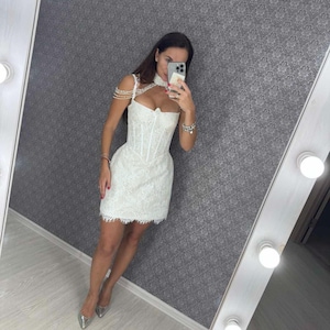 K&ouml;nnte beinhalten: Wei&szlig;es Mini-Kleid aus Spitze mit einem Korsett-Oberteil und Perlenriemendetails. Das Kleid hat einen gewellten Saum und wird mit silbernen Abs&auml;tzen getragen. Das Kleid spiegelt sich in einem beleuchteten Spiegel.
