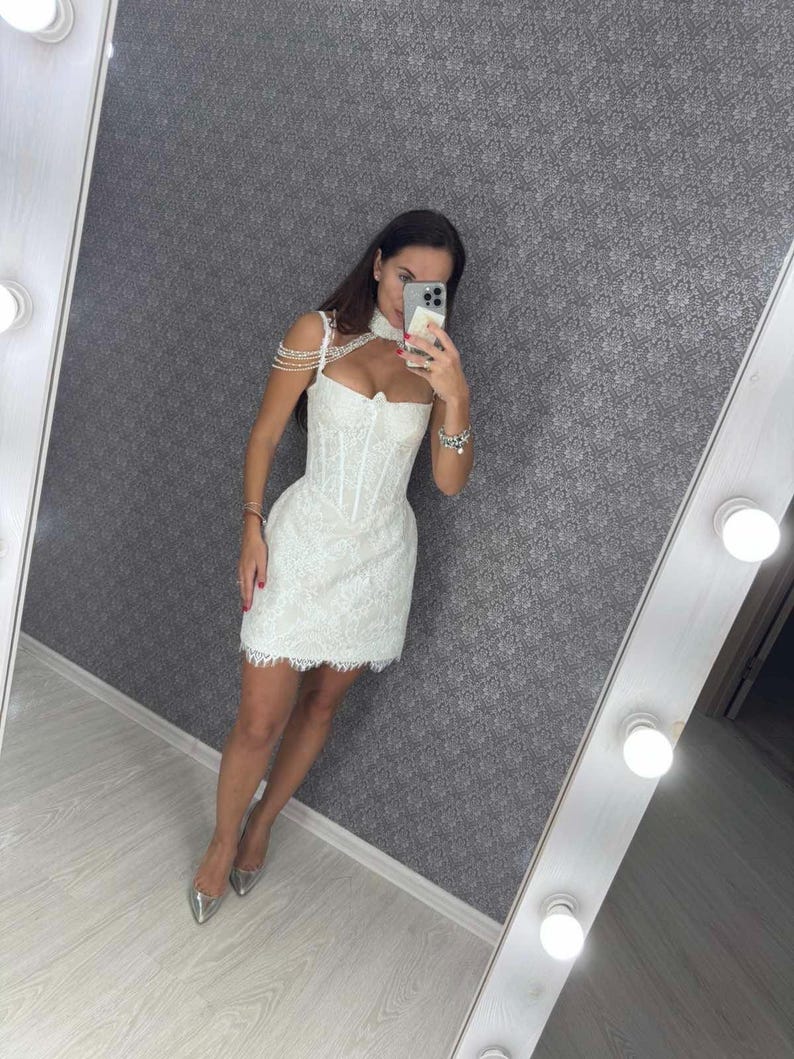 K&ouml;nnte beinhalten: Wei&szlig;es Mini-Kleid aus Spitze mit einem Korsett-Oberteil und Perlenriemendetails. Das Kleid hat einen gewellten Saum und wird mit silbernen Abs&auml;tzen getragen. Das Kleid spiegelt sich in einem beleuchteten Spiegel.