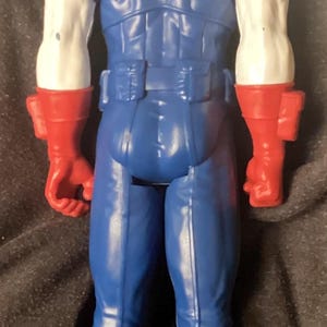 May include: A blue action figure of a superhero, viewed from the back. The figure has white arms with red gloves and red boots. The figure's back has a star design.