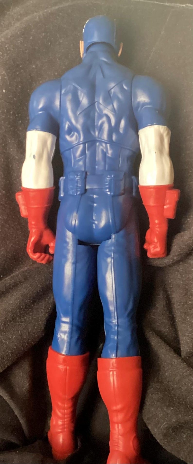 May include: A blue action figure of a superhero, viewed from the back. The figure has white arms with red gloves and red boots. The figure's back has a star design.