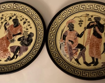 May include: Two decorative plates with black rims and a cream-colored center. Each plate features a Greek-style scene with figures in orange, purple, and black, surrounded by a geometric border. The plates are likely for display.
