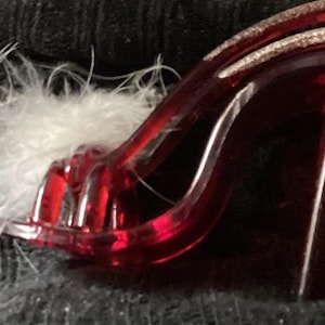 May include: A red high-heeled shoe with a clear, glossy finish. The shoe features a decorative white feather accent and a silver glitter detail on the straps. The heel is a solid, rectangular shape.
