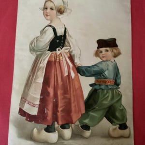 May include: An antique postcard featuring a painting of two children in traditional Dutch clothing. The girl wears a white cap, a black bodice, and a red skirt. The boy wears a blue shirt, green pants, and a brown cap. Both children are wearing wooden shoes.