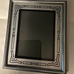 May include: A rectangular silver picture frame with a black and silver geometric design around the inner edges. The frame has a glass insert and is suitable for displaying photographs or artwork. The frame is on a white surface.