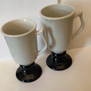 May include: Two white ceramic coffee mugs with black bases. The mugs have a footed design and are perfect for serving coffee or tea.