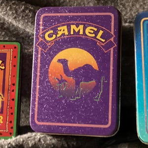 May include: Three vintage Camel cigarette tin boxes. The boxes feature the Camel logo with a camel silhouette and text. One box reads "Beach Breezer", another has a sunset design, and the third says "Joe's Place".