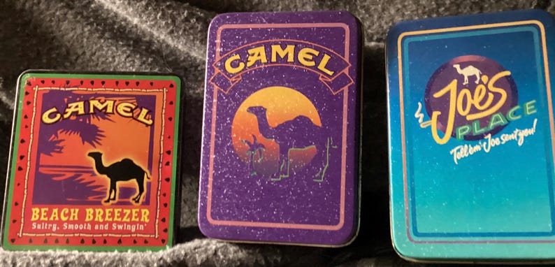 May include: Three vintage Camel cigarette tin boxes. The boxes feature the Camel logo with a camel silhouette and text. One box reads "Beach Breezer", another has a sunset design, and the third says "Joe's Place".