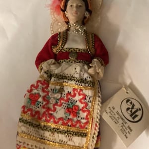 May include: A detailed doll in traditional attire, featuring a red jacket with gold trim, a white blouse, and a patterned skirt with red floral embroidery. The doll has a decorative headpiece and a tag attached.