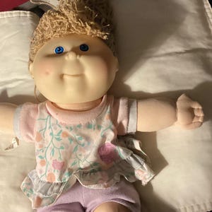 May include: A vintage doll with a soft, vinyl face, blue eyes, and a light brown yarn hairstyle. The doll wears a pink top with a floral pattern and lilac shorts. The doll's arms are outstretched.
