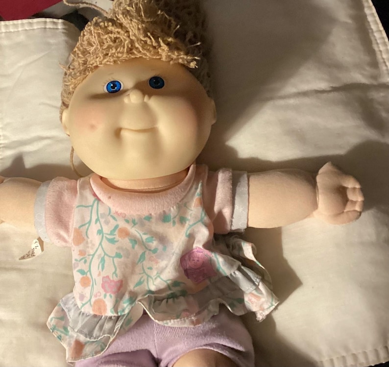 May include: A vintage doll with a soft, vinyl face, blue eyes, and a light brown yarn hairstyle. The doll wears a pink top with a floral pattern and lilac shorts. The doll's arms are outstretched.