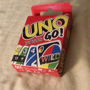 May include: A red box of UNO GO! pocket-sized playing cards. The box features the game's logo in yellow and black, with card illustrations showing numbers and the word "WILD". The text "Pocket-Sized Cards For On-The-Go Play!" is also visible.