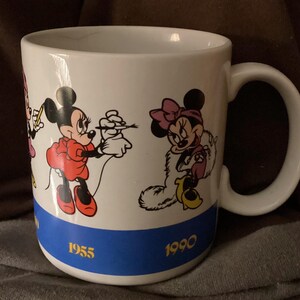 May include: White ceramic mug featuring Minnie Mouse in various poses. The mug has a blue band at the bottom with the years 1955 and 1990. The handle is white and the mug is likely for hot beverages.