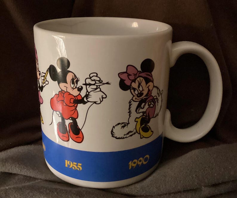 May include: White ceramic mug featuring Minnie Mouse in various poses. The mug has a blue band at the bottom with the years 1955 and 1990. The handle is white and the mug is likely for hot beverages.