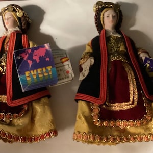 May include: Two porcelain dolls in traditional attire. Each doll wears a gold and red dress with a black overcoat, a decorative headpiece, and holds a tag with the word "EVELT".