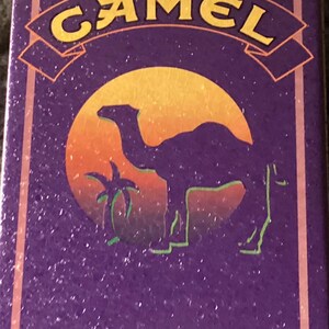 May include: Rectangular purple tin with the word "CAMEL" in yellow lettering. The tin features a camel silhouette in front of a sunset with a palm tree. The border is a lighter shade of purple.