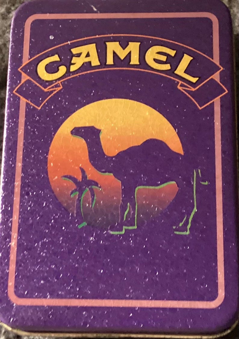 May include: Rectangular purple tin with the word "CAMEL" in yellow lettering. The tin features a camel silhouette in front of a sunset with a palm tree. The border is a lighter shade of purple.