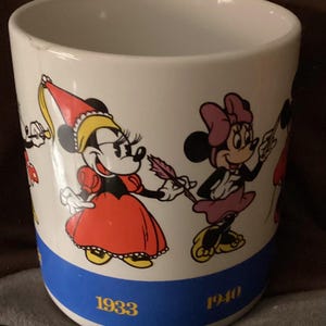 May include: White ceramic mug featuring Minnie Mouse in various outfits. The mug has a blue band with the years 1933 and 1940 printed on it. The mug has a few small chips on the rim.